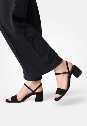 Voeten in zwarte sandalen met blokhak en enkelbandjes, gecombineerd met zwarte cropped wijde broekspijpen tegen een effen achtergrond.