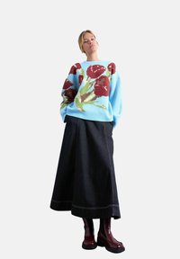Jeune femme portant un pull bleu clair avec de grands motifs floraux rouges, une longue jupe noire et des bottines brillantes rouge foncé, debout les mains dans les poches.