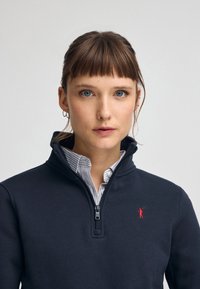 Felpa zip-up blu navy realizzata in morbido materiale, con un accento ricamato rosso e una camicia con colletto a righe bianche sotto.