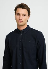 Mørkeblå langermet skjorte laget av teksturert stoff, med krage som kan kneppes ned, frontlomme og svarte knapper.