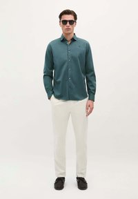 Cămașă butonată cu manșete lungi, de culoare verde închis, pantaloni de culoare deschisă, pantofi negri și ochelari de soare; textură netedă, design mulat, stil minimalist.