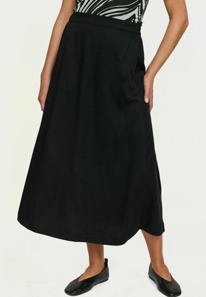 Femme portant une jupe midi noire associée à des ballerines noires et un haut à imprimé zèbre, debout devant un fond clair uni.