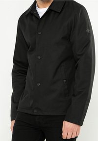 Czarna kurtka z teksturowanej tkaniny, zapinana na guziki, z wyprofilowanym kołnierzem i bocznymi kieszeniami, z naszywką logo na rękawie.