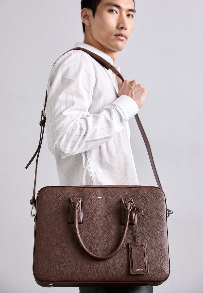 Vyras su balta juostuota marškinėliais, nešantis rudą odinę portfelį su petine juosta ir rankenomis, prieš pilką foną.