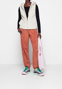 Krémová umělá kožešinová vesta s kapucí, černé triko s dlouhými rukávy, oranžové tepláky s logem Nike a růžov�é tenisky se zelenými akcenty. Drží růžovou tašku.