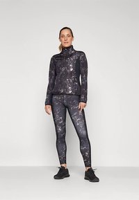 Vrouw die staat in een zwart en grijs gemarmerde sportjack en leggings met zwarte sneakers tegen een vlakke lichte achtergrond.
