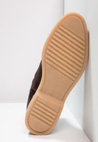 Zapato de ante marrón con suela de goma, que presenta estrías horizontales para un mejor agarre. La suela es de un tono marrón claro, mostrando un diseño limpio y aerodinámico.