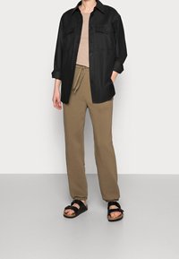 Camisa negra de botones con dos bolsillos en el pecho, camiseta beige ajustada, pantalones de chándal color caqui con cordón y sandalias negras con hebillas.
