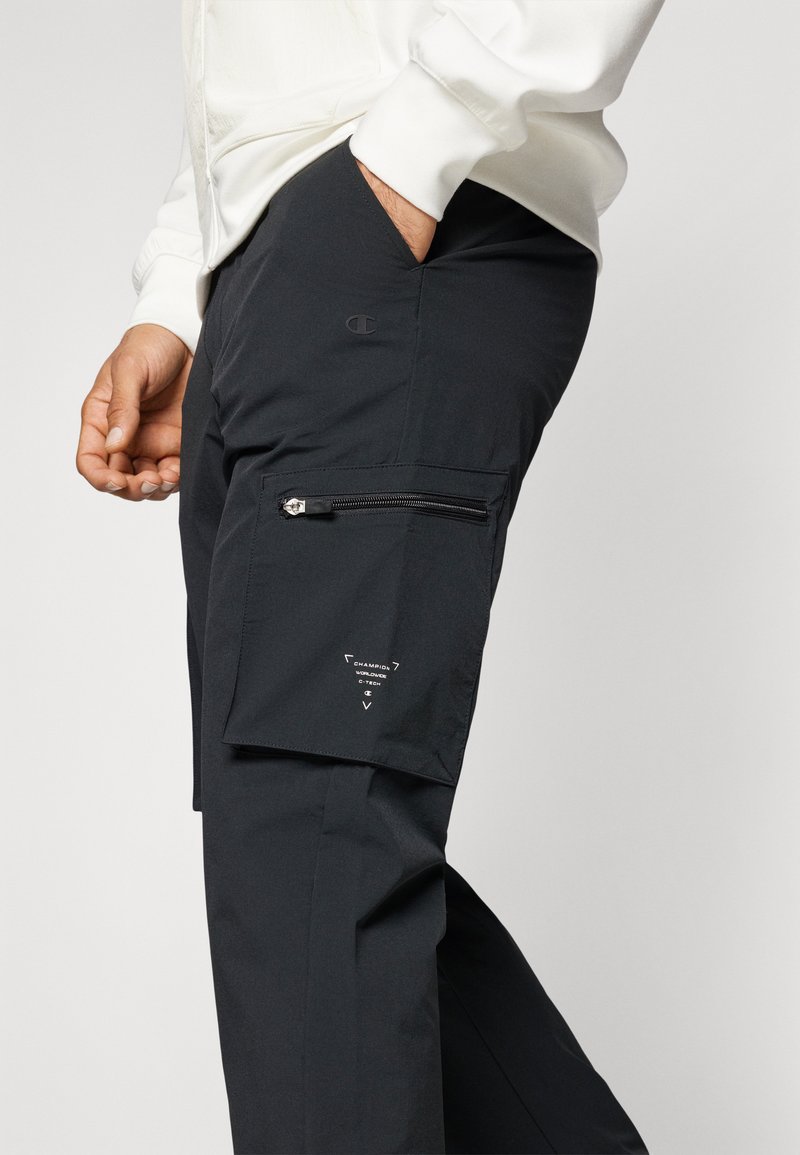 Pantalons cargo noirs en tissu léger, avec une poche latérale zippée et un détail du logo sur la cuisse. Texture lisse avec un design ajusté.