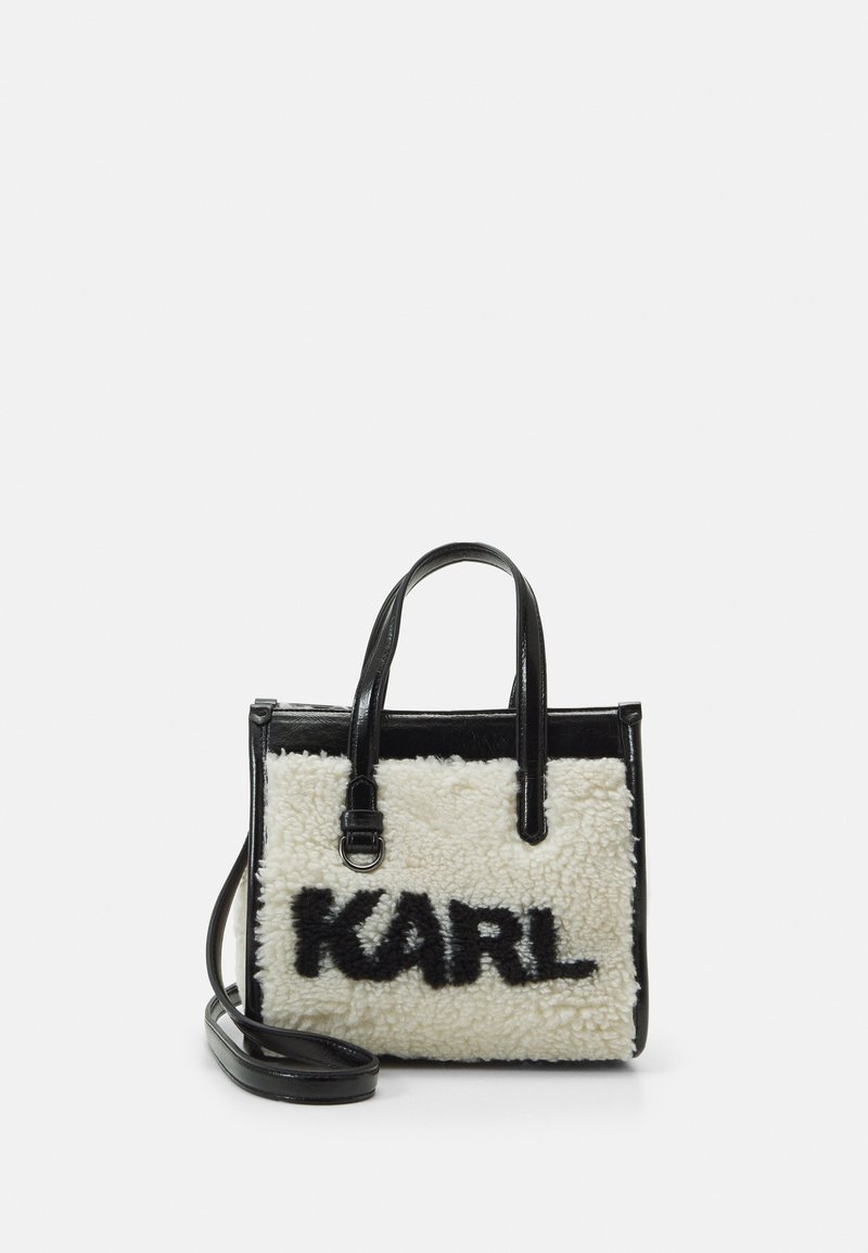 Svart och vit tygväska med en plyschig, fluffig textur som har ordet "KARL" i svart. Den har två handtag upptill och en justerbar axelrem.