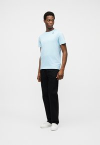 Άνδρας όρθιος, φορώντας ανοιχτό μπλε κοντομάνικο μπλουζάκι, μαύρο παντελόνι και λευκά αθλητικά παπούτσια, μπροστά σε απλό λευκό φόντο.