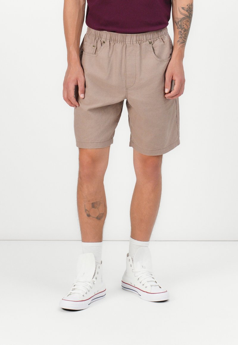 Pantalones cortos beige con cintura elástica, dos bolsillos frontales y corte relajado, combinados con zapatillas altas blancas y calcetines blancos.