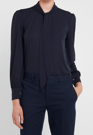 Vrouw die een marineblauwe blouse met een strik aan de voorkant en gouden knoopmanchetten draagt, gecombineerd met marineblauwe, getailleerde broek, met één hand in de zak tegen een effen achtergrond.