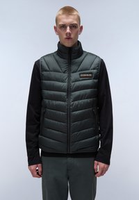 Grön pufferväst med quiltat mönster, dragkedja, hög krage och logotyp-patch. Lager på en svart långärmad skjorta och gråbyxor.