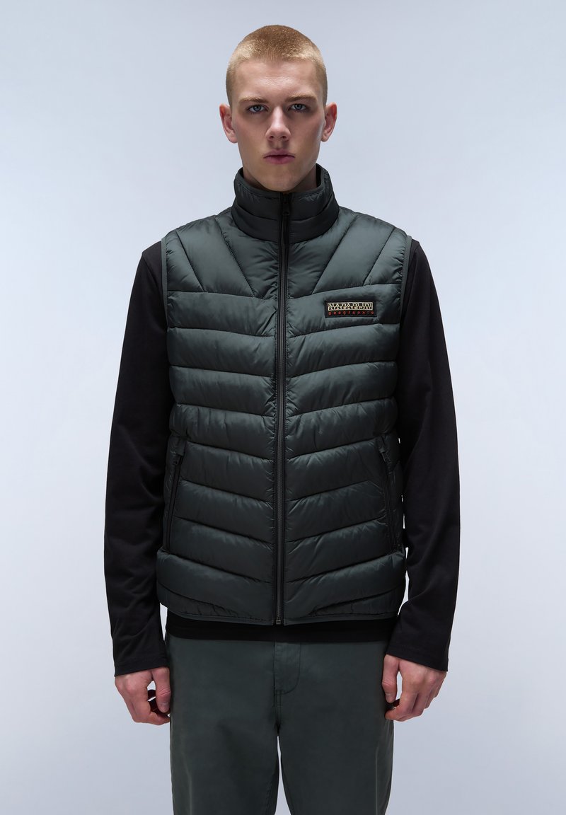 Grön pufferväst med quiltat mönster, dragkedja, hög krage och logotyp-patch. Lager på en svart långärmad skjorta och gråbyxor.