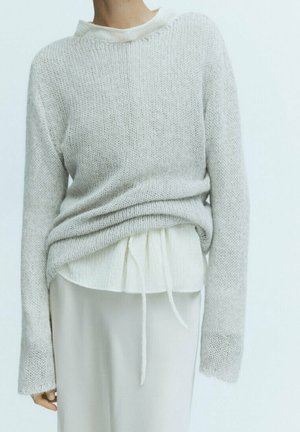 Person iført en lysegrå strikket sweater over en hvid bluse med snøre i taljen og et hvidt skørt mod en ensfarvet baggrund.