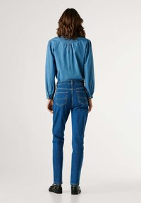 Denimová košile s nabíranými rukávy; modré úzké džíny s vysokým pasem a zadními kapsami; černé ploché boty; neutrální pozadí.