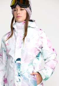 Witte waterdichte snowjack met meerkleurige bloemenpatronen in pastelkleuren, hoge kraag, ritszakken en verstelbare manchetten.