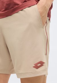 Άτομο που φοράει μπεζ σορτς με το χέρι στην τσέπη, μικρό κόκκινο λογότυπο κοντά στο τελείωμα και ένα κόκκινο πουκάμισο που διακρίνεται μερικώς πάνω από τη μέση.