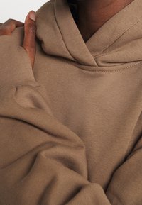 Persona dalla pelle scura che indossa una felpa marrone con cappuccio, mano appoggiata sulla spalla opposta, tessuto leggermente stropicciato vicino al gomito.