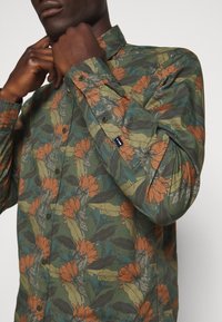 Homme ajustant le col d’une chemise verte à manches longues avec imprimé floral orange et bleu et poignets boutonnés sur fond blanc.