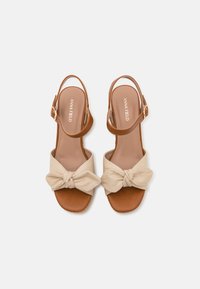 Brown leather sandals with a beige fabric bow accent, open-toe design, and an adjustable ankle strap. Smooth texture and casual style.
