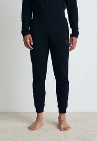 Pantalons de survêtement noirs en tissu doux, avec un design ajusté, des chevilles côtelées et un petit patch de logo rouge sur la cuisse gauche.