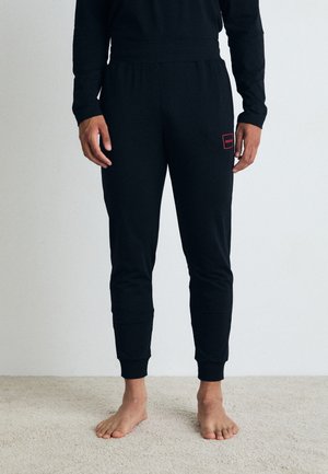 Pantalons de survêtement noirs en tissu doux, avec un design ajusté, des chevilles côtelées et un petit patch de logo rouge sur la cuisse gauche.