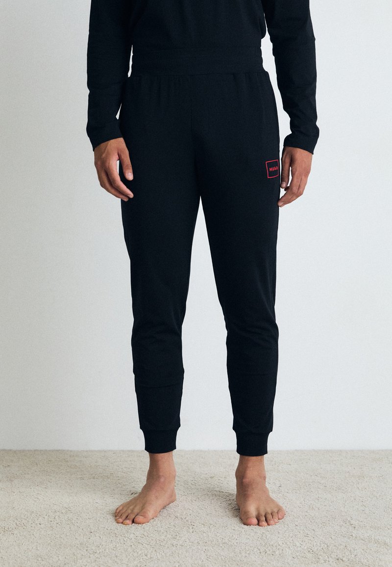 Pantalons de survêtement noirs en tissu doux, avec un design ajusté, des chevilles côtelées et un petit patch de logo rouge sur la cuisse gauche.