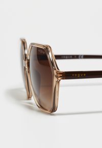 Brown-tinted oversized sunglasses with a transparent, light brown frame. Features gold "vogue" logo on the temple and sleek, angular design.