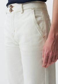 Pantalon taille haute beige en tissu texturé, doté de deux boutons marron et de poches latérales, rehaussé de coutures contrastantes.