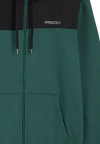 Sudadera con capucha y cremallera confeccionada en tela verde con un panel superior negro. Cuenta con bolsillos delanteros, cordones ajustables y un logotipo "FREDDY".