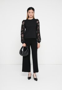 Top negro con mangas largas transparentes y texturizadas, combinado con pantalones anchos negros. Accesorios incluyen un pequeño bolso negro y zapatos planos de punta.