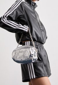 Metallisch zilveren handtas met kettingriem, met een reliëf logo "Juicy Couture", vastgehouden door een persoon in een zwarte sportoutfit met witte strepen.