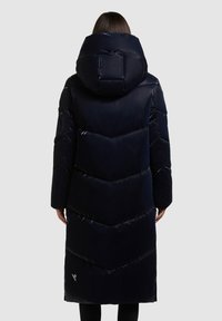 Dlouhý navy blue puffer kabát s velkou kapucí, který má chevronový design, lesklou texturu a detail loga na boku.