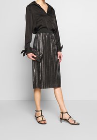 Blouse en satin noir avec col, jupe plissée argentée métallique, et sandales noires à brides, tenant une pochette noire texturée.