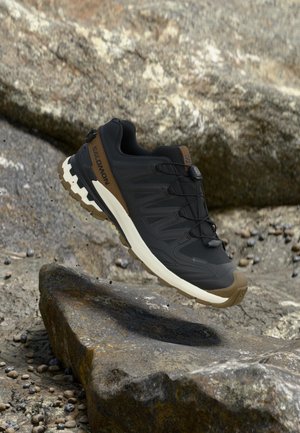 Melns un brūns Salomon pārgājienu apavu pāris ar raupju zoli, kas virs klints ainavas.