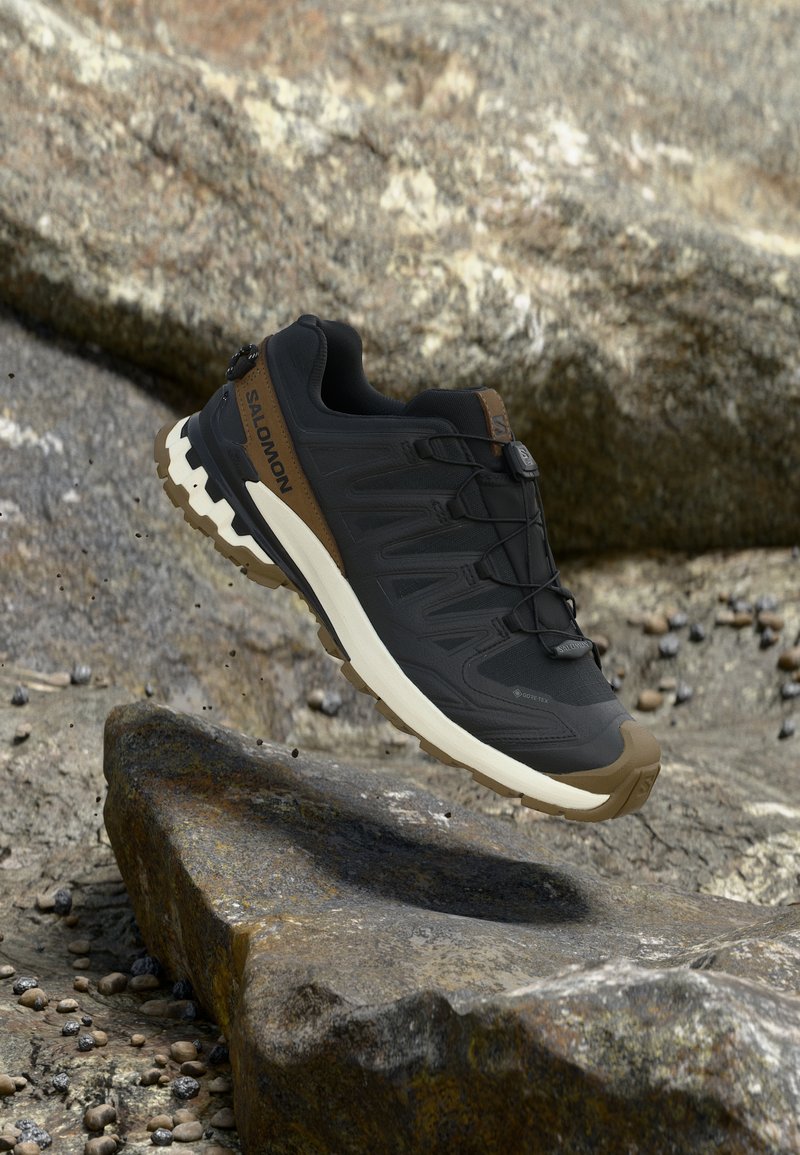 Μαύρο και καφέ πεζοπορικό παπούτσι Salomon με ανθεκτική σόλα, αιωρούμενο πάνω από πετρώδες έδαφος.