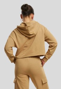 Sudadera recortada de tela marrón claro con una textura suave, que cuenta con capucha y mangas largas, combinada con pantalones a juego que tienen bolsillos laterales.