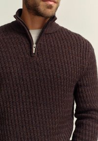 Brauner gerippter Pullover mit halbem Reißverschluss, strukturiertem Strick und taillierter Passform. Der Stoff wirkt weich und warm.