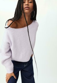 Ljust lila, ribbat stickad tröja med off-the-shoulder design och långa ärmar, kombinerat med mörkblå denimbyxor.