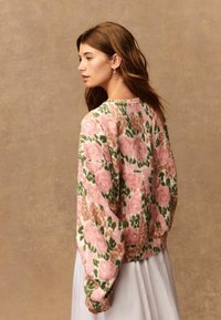 Junge Frau mit welligem braunem Haar trägt einen blumigen Pullover in Rosa, Grün und Beige sowie einen weißen Rock und steht nach links gegen einen beigen Hintergrund.