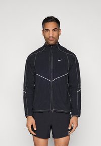 Črna Nike vetrovka z odsevnimi šivi, visokim ovratnikom in zadrgo spredaj. Nošena s črnimi kratkimi hlačami. Ravna zasnova, lahka tkanina.