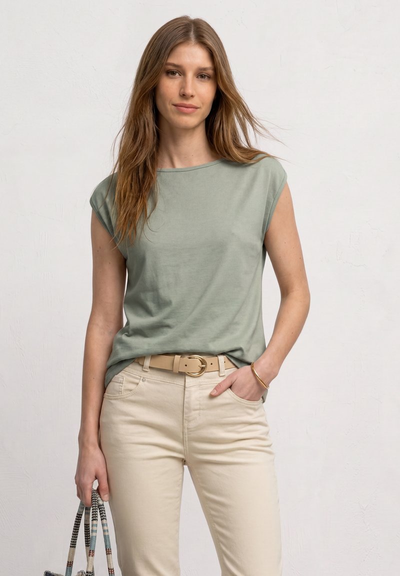 Femme aux longs cheveux bruns portant un haut sans manches vert sauge et un pantalon beige, tenant un sac à main tressé avec une main dans la poche.