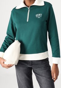 Suéter verde con cuello blanco y cierre de cremallera. Presenta puños y dobladillo acanalados, logotipo de la marca en el pecho y un acabado de algodón suave.