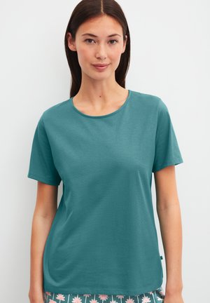 Jonge vrouw met steil donker haar, gekleed in een donkergroene T-shirt met korte mouwen en een broek met bloemmotief, staand tegen een witte achtergrond.