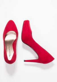 Červená semišová bota na vysokém podpatku s špičatou špičkou a hladkým kovovým vnitřkem. Má štíhlý podpatek a polstrovanou podrážku.