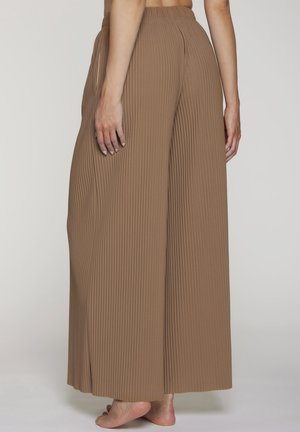 Woman barefoot wearing high-waisted, wide-leg, ribbed brown pants, shown from the back against a plain light background.