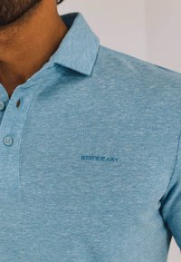 Lichtblauwe polo van textuurstof, met een kraag, twee knopen en een klein logo geborduurd op de borst.
