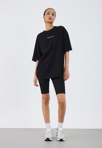 Schwarzes, oversized T-Shirt mit weißem Text, kombiniert mit schwarzen Radshorts. Getragen mit weißen Socken und grauen Sneakers vor einem schlichten Hintergrund.